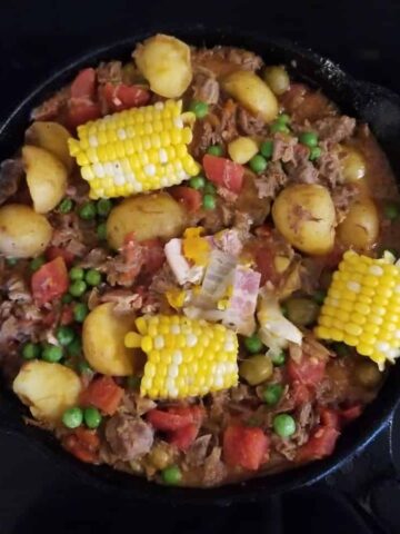 Puerto Rican Beef Stew with baby golden potatoes, corn on the cob,green peas, stewed tomatoes, and bacon in a brown sauce in a black cast iron skillet
