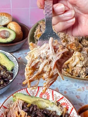 A fork full of Pernil with a large serving bowl, avocados, dinner rolls, and small serving bowls with meat on the background.