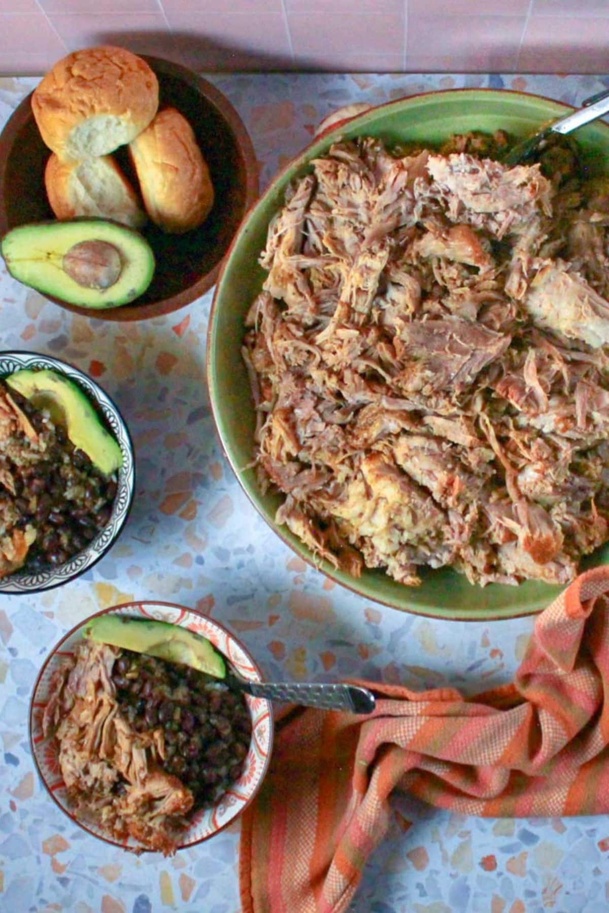 Table setting of a large green bowl full of pernil, with a small bowl of bread rolls to the left and two small bowls full of rice, black beans, pork, and avocado.