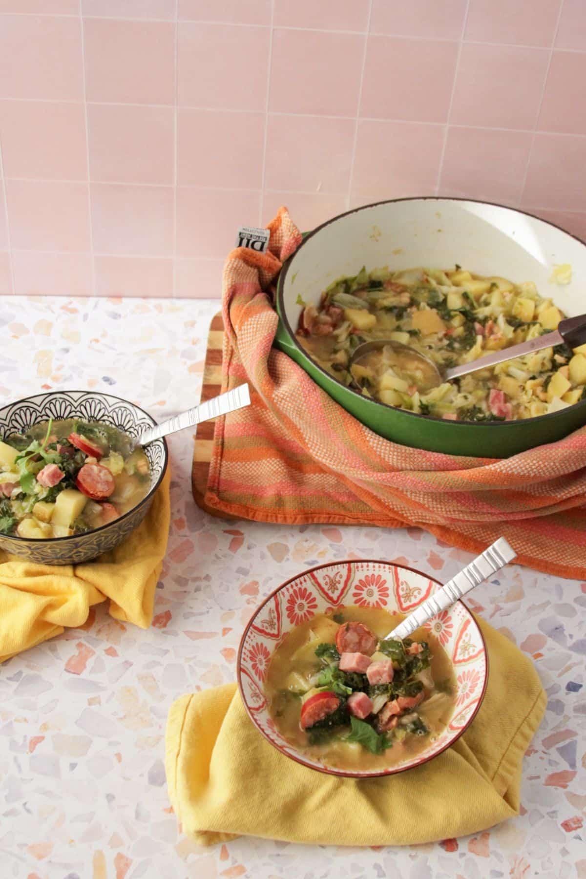 Two bowls of Caldo Gallego served sitting atop of two yellow cloth napkins. Large green dutch oven with a kitchen towel wrapped around it sits in the background.