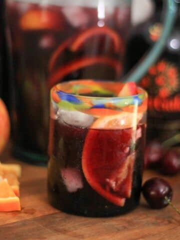 A glass of red sangria with ice and fruit garnish surrounded by cut fruits, the pitcher of sangria, and bottles of wine and liqueur.