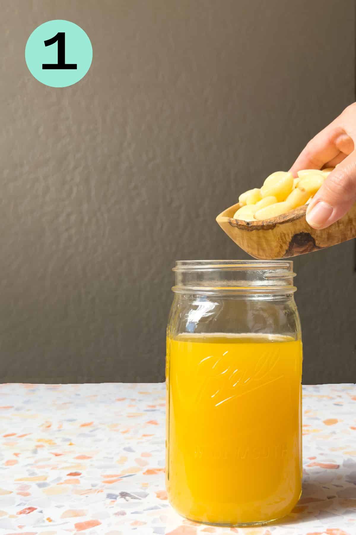 A mason jar filled with orange juice on a terrazo top table. Garlic cloves are being added to the jar.