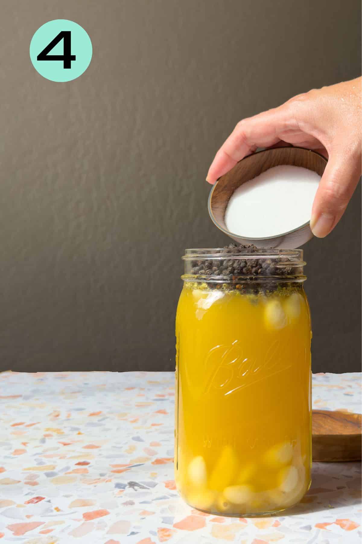 A mason jar filled with orange juice, oregano, peppercorns, and garlic on a terrazo top table. Kosher salt is being added to the jar.