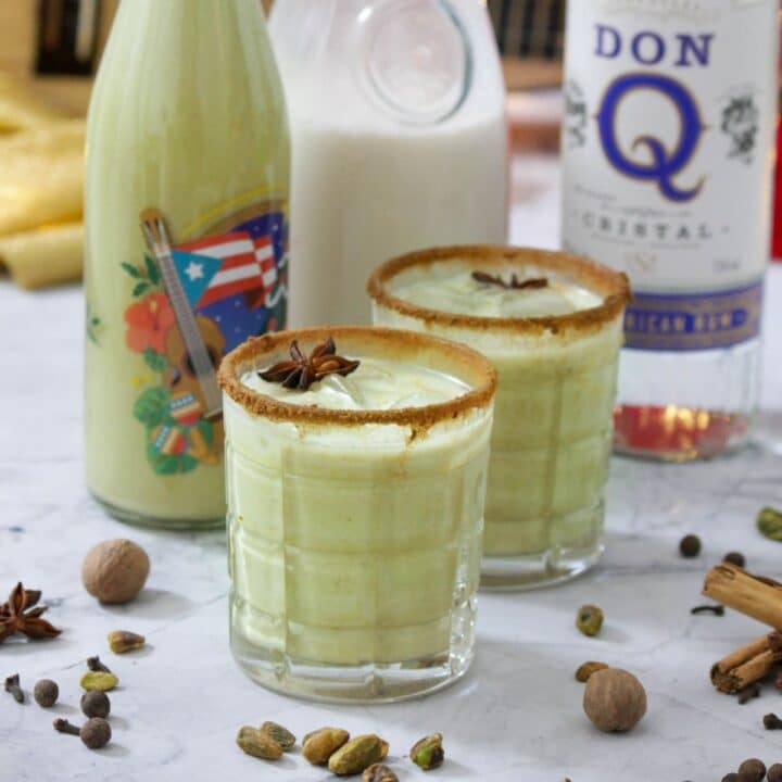 Two rock glassed filled with pistachio coquito and decorated with cinnamon sugar on the rims. A Puerto Rican Christmas decorated bottle sits behind them along with a bottle of regular coquito and a bottle of rum.