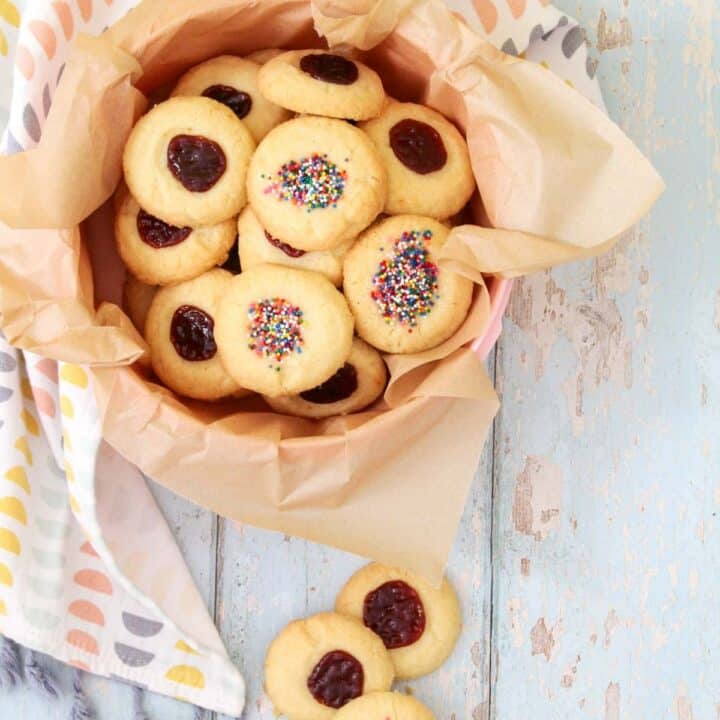 Tupperware cookie container lined with brown parchment paper and full of polvorones on a table with a kitchen towel around the container and some loose cookies in front of it.