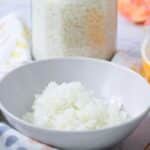 Bowl of cooked white rice with a jar of uncooked rice behind it.