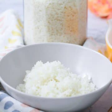 Bowl of cooked white rice with a jar of uncooked rice behind it.