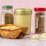 Small mason jar filled with Puerto Rican Garlic Paste with a jar of oregano and a jar of coriander seeds at either side of it. A small wooden bowl with garlic paste sits in front of the mason jar with garlic paste as well as a few garlic cloves.