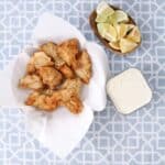 Bowl lined with paper towels and filled with fried mero chunks. A small bowl with cut lemons and a small bowl with tartar sauce.