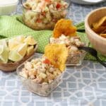 An individual serving of ceviche with a plantain toston stuck on the back sits behind a bowl of lemons and limes and a larger bowl of ceviche.