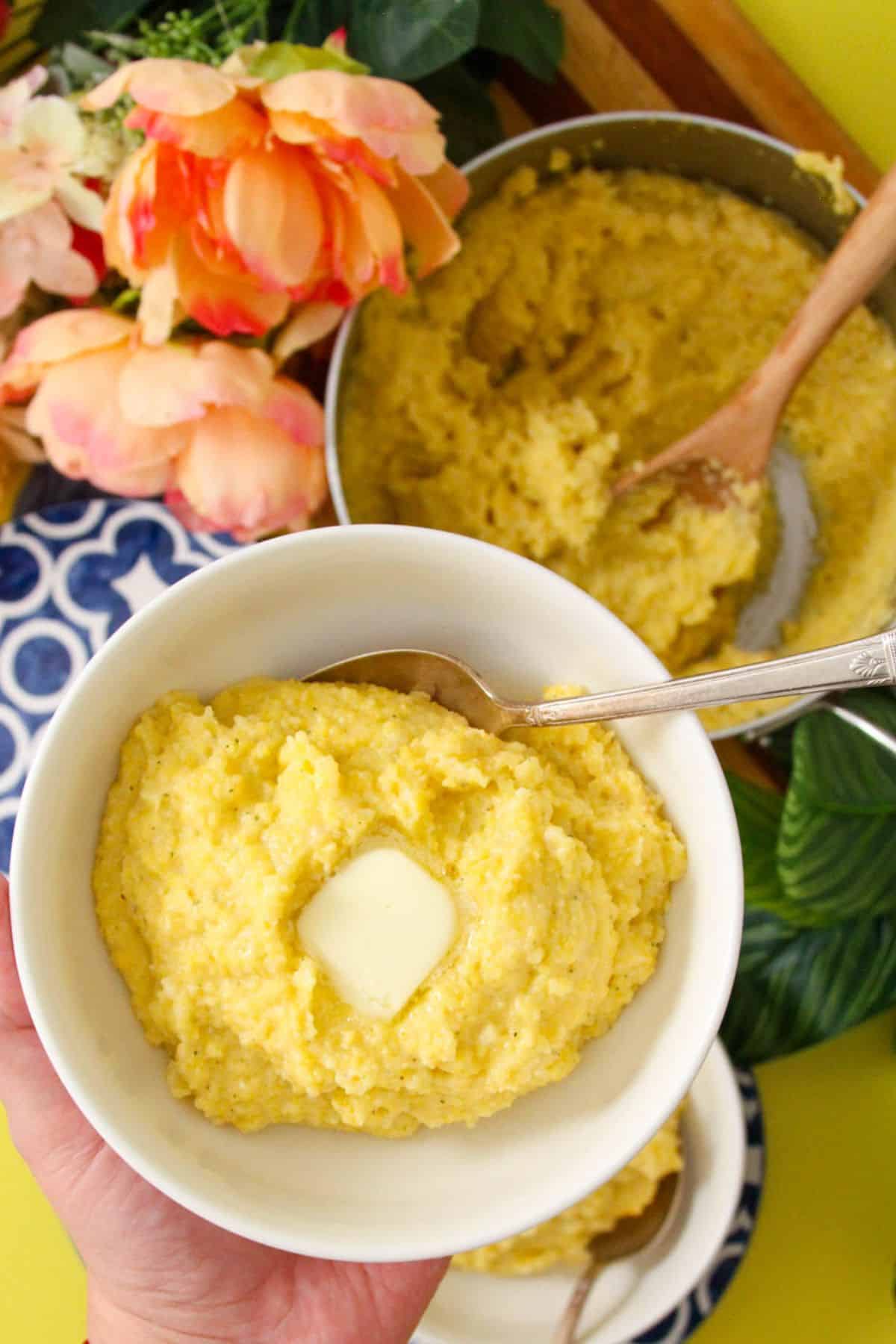 A hand holding a white bowl of funche topped with a dab of butter.