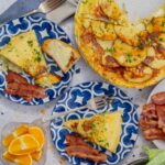 A slice of Spanish tortilla (Tortilla Española) made with eggs and sliced potatoes is served on a blue-patterned plate alongside crispy bacon. The full tortilla is visible in the background, garnished with chopped parsley. Toast with melted cheese, extra bacon, and a basket of sliced bread are also seen on the table.