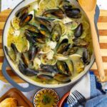 A large Dutch oven filled with steamed mussels and clams in a light sofrito and white wine broth, garnished with herbs and red pepper flakes.