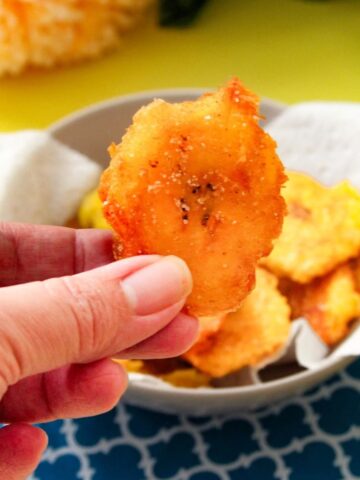 A hand holding a smashed plantain. The rest of the plantains are in a towel-lined bowl sits behind it.
