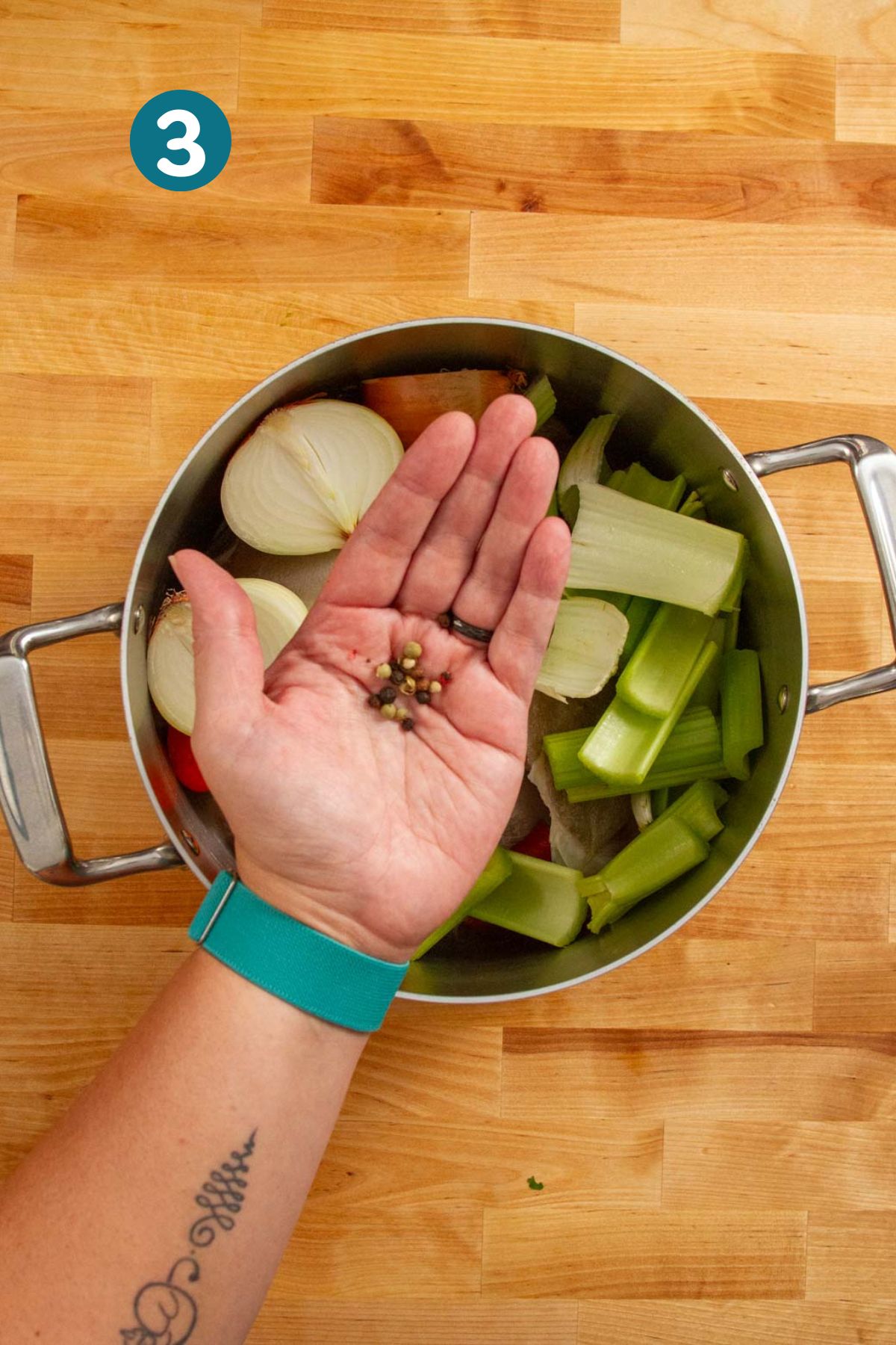 A hand holding whole peppercorns above the pot, about to add them to the broth.