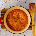 Overhead view of Puerto Rican Crab Rice (Arroz con Jueyes) in a red Dutch oven, showing rice simmered with crab meat, olives, red peppers, and sofrito. A wooden slotted spatula rests beside the pot on a terrazzo countertop.