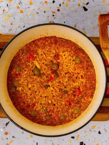 Overhead view of Puerto Rican Crab Rice (Arroz con Jueyes) in a red Dutch oven, showing rice simmered with crab meat, olives, red peppers, and sofrito. A wooden slotted spatula rests beside the pot on a terrazzo countertop.