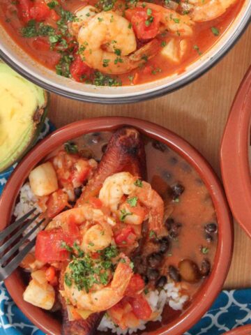 Close-up of shrimp and scallop stew over white rice, black beans, and fried sweet plantain, with a side of avocado and extra stew in the background.