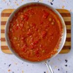 Overhead view of a skillet filled with Mojo Isleño, a chunky Puerto Rican tomato sauce with olives, onions, and garlic, simmering on a striped wood board.