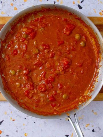 Overhead view of a skillet filled with Mojo Isleño, a chunky Puerto Rican tomato sauce with olives, onions, and garlic, simmering on a striped wood board.