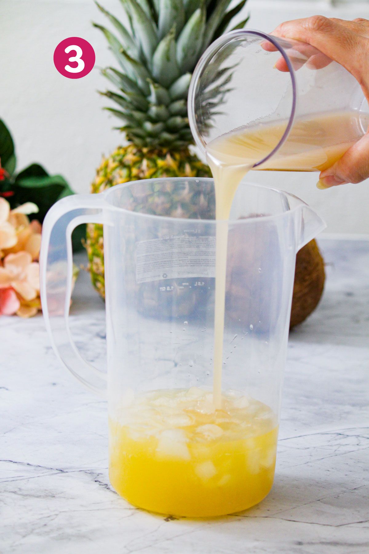 Cream of coconut is being poured into a pitcher with pineapple juice and ice.