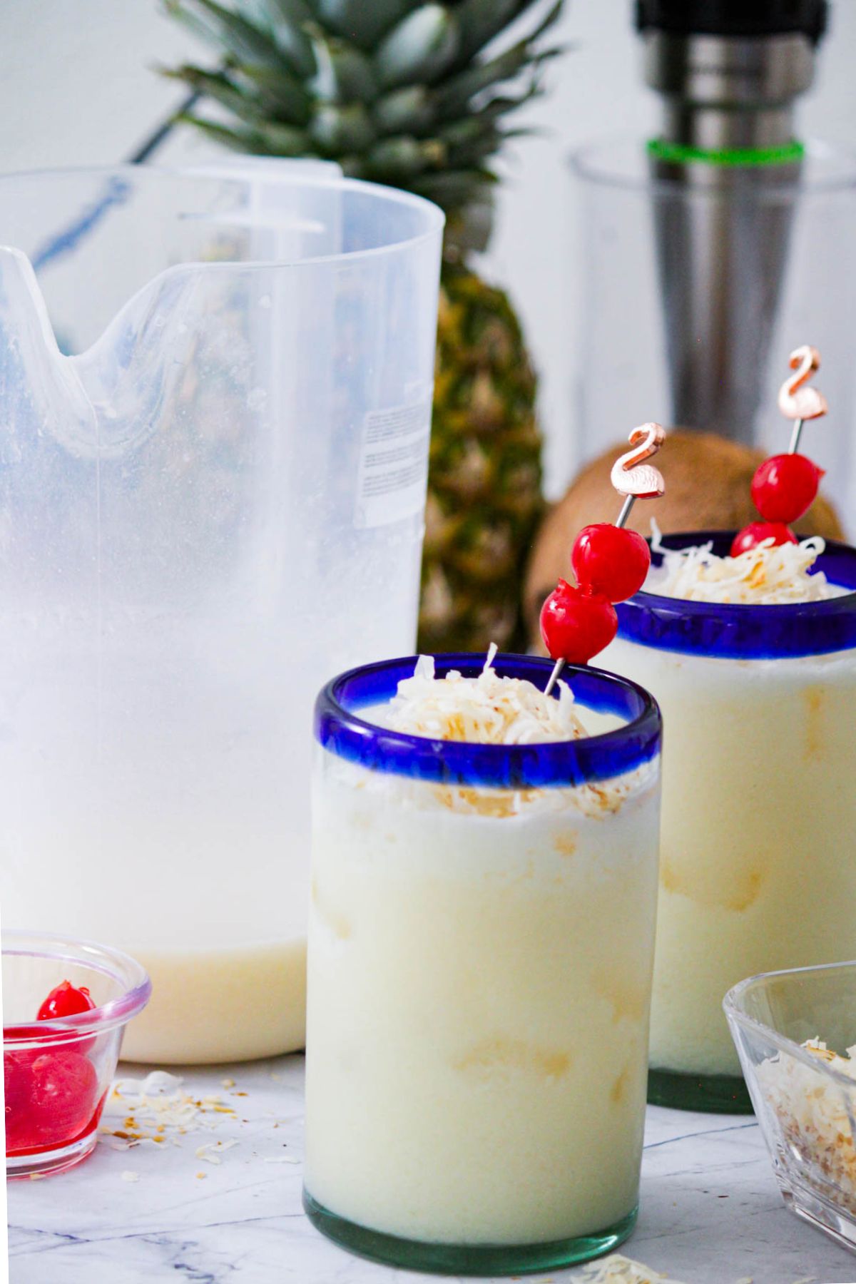 Close-up of piña colada garnished with toasted coconut and cherries in a blue-rimmed glass with pitcher in background.