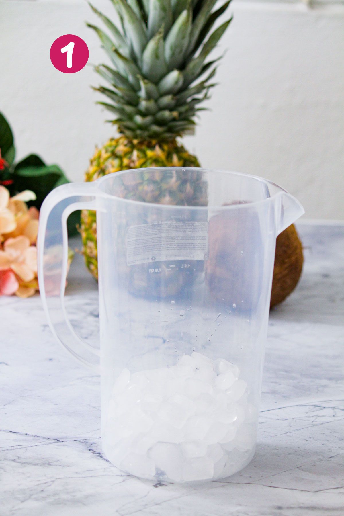 A large plastic pitcher, partially filled with ice cubes, ready to start the piña colada mix, with a pineapple in the background.
