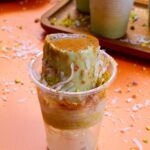 Close-up of a creamy pistachio limber topped with cinnamon, shredded coconut, and chopped pistachios, partially popped out of its plastic cup on an orange surface.