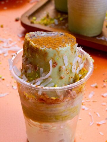 Close-up of a creamy pistachio limber topped with cinnamon, shredded coconut, and chopped pistachios, partially popped out of its plastic cup on an orange surface.