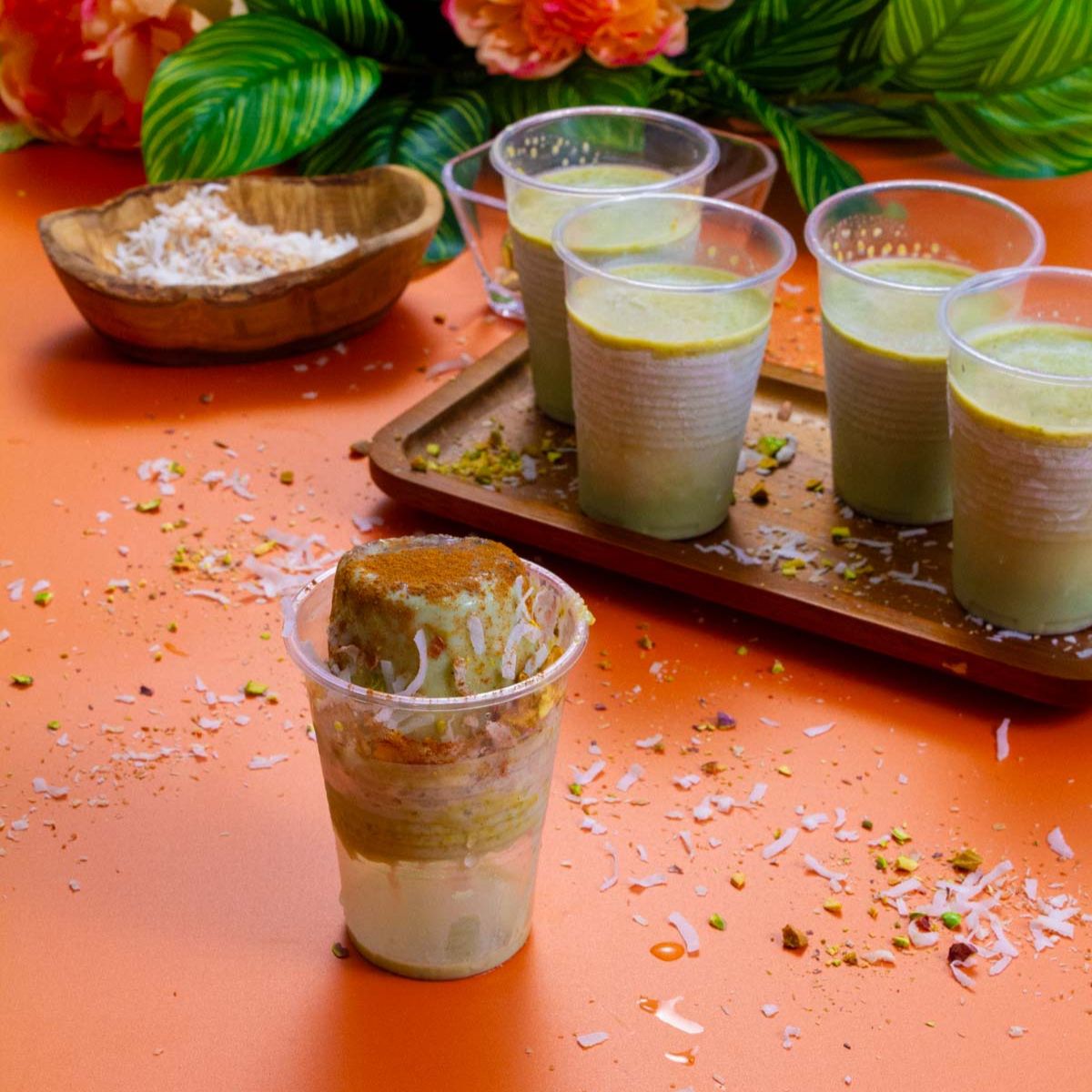 Pistachio limbers in clear plastic cups, one popped and garnished in front, with others chilling on a tray in the background. Surrounded by coconut flakes and chopped pistachios on an orange table.