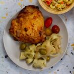 Close-up of a roasted chicken thigh served with guineos en escabeche, cherry tomatoes, and a side of macaroni salad partially visible.