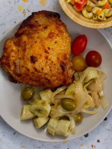 Close-up of a roasted chicken thigh served with guineos en escabeche, cherry tomatoes, and a side of macaroni salad partially visible.
