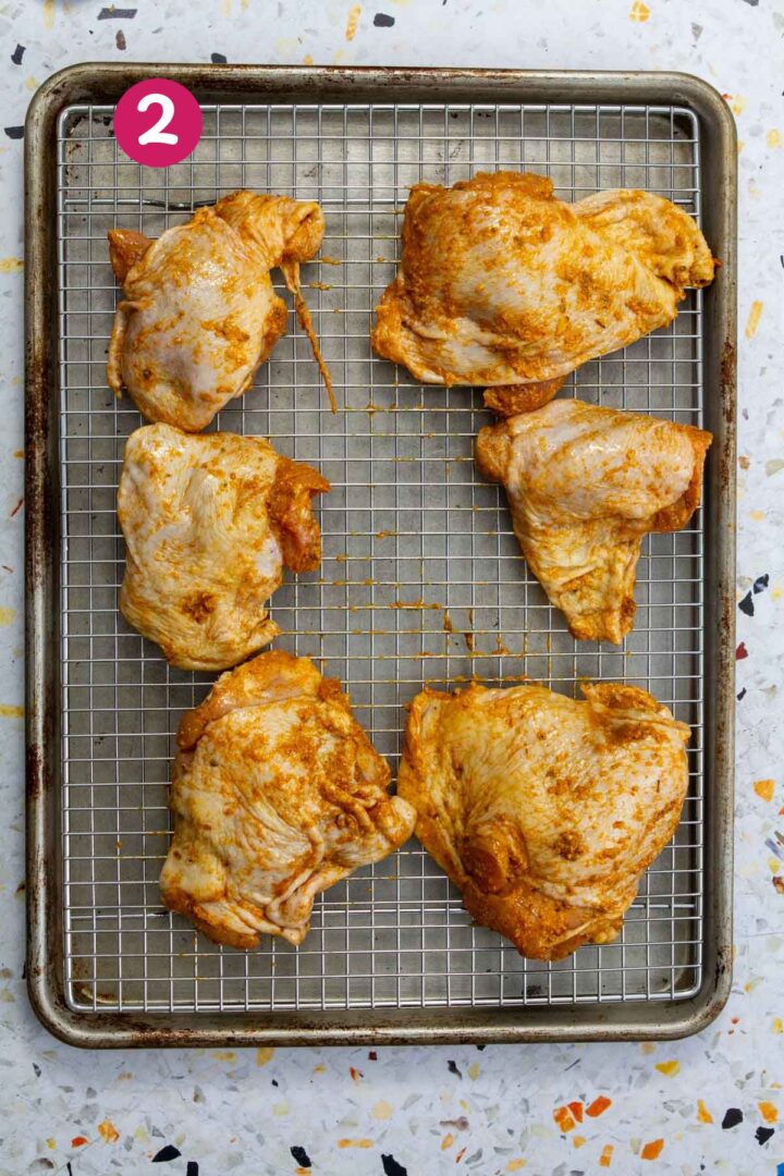 Seasoned chicken thighs arranged on a wire rack over a sheet pan, ready to go into the oven.