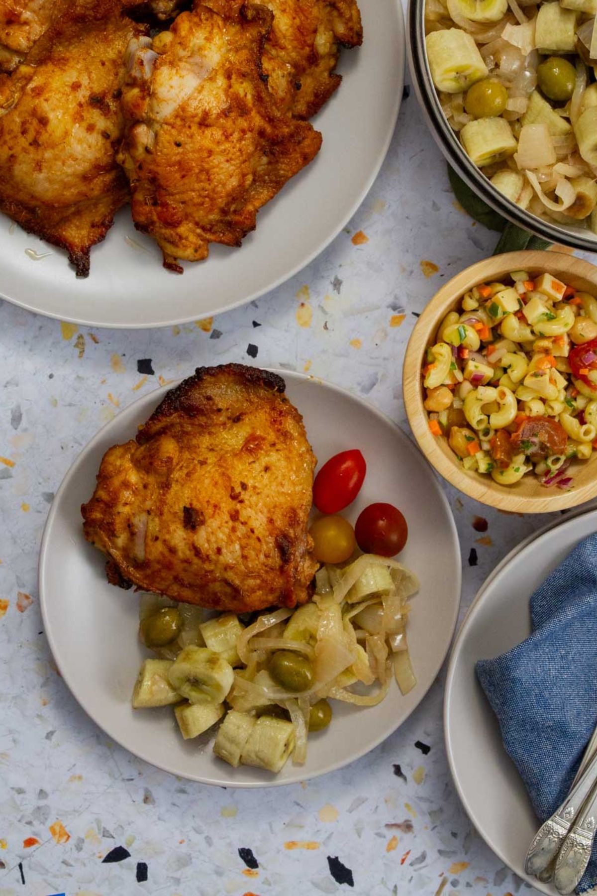 Top-down view of a plate with one roasted chicken thigh, guineos en escabeche, and cherry tomatoes, next to bowls of macaroni salad and additional chicken.