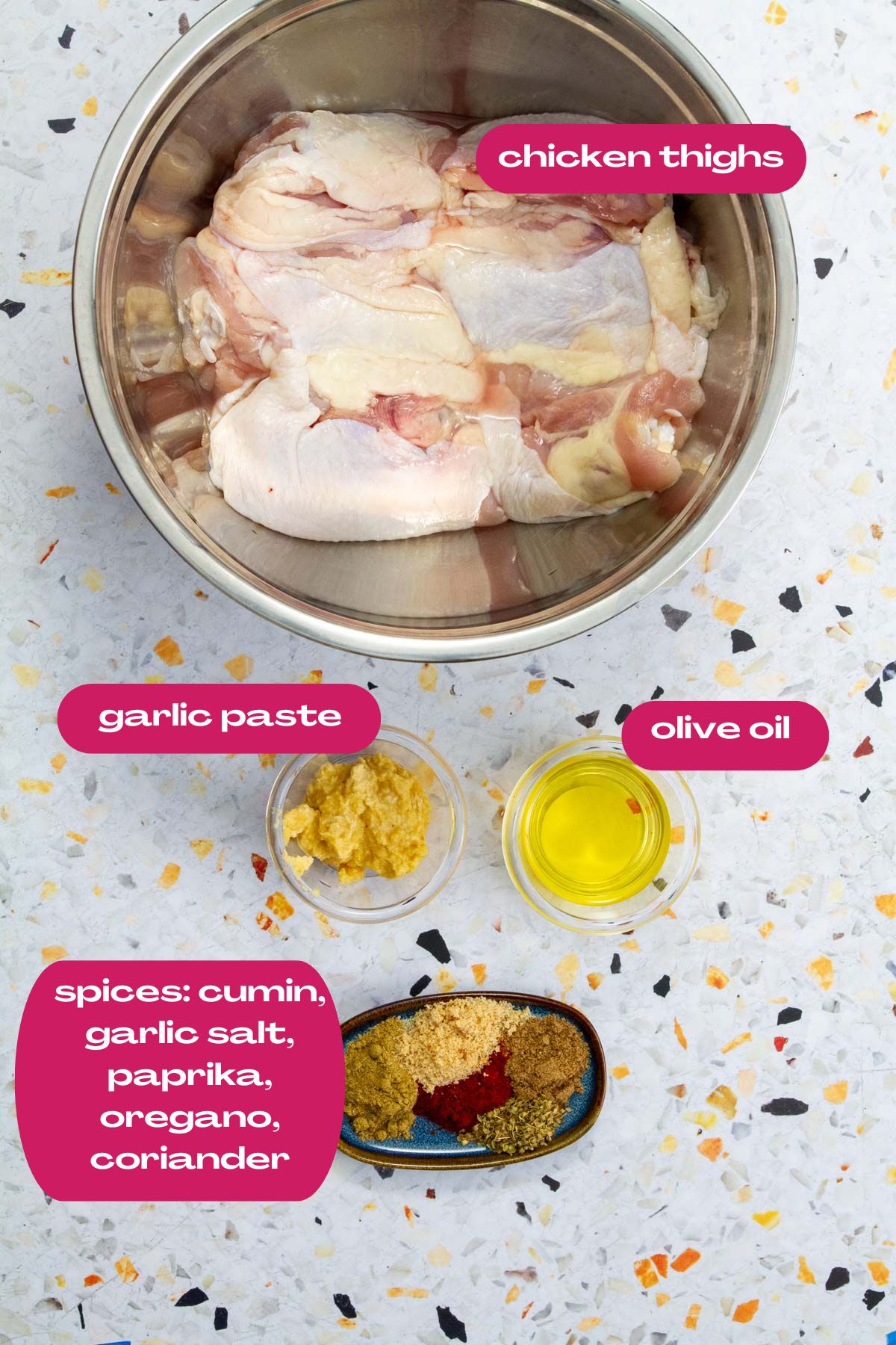 Raw chicken thighs in a metal bowl surrounded by labeled ingredients: garlic paste, olive oil, and a small dish of ground spices including cumin, garlic salt, paprika, oregano, and coriander.