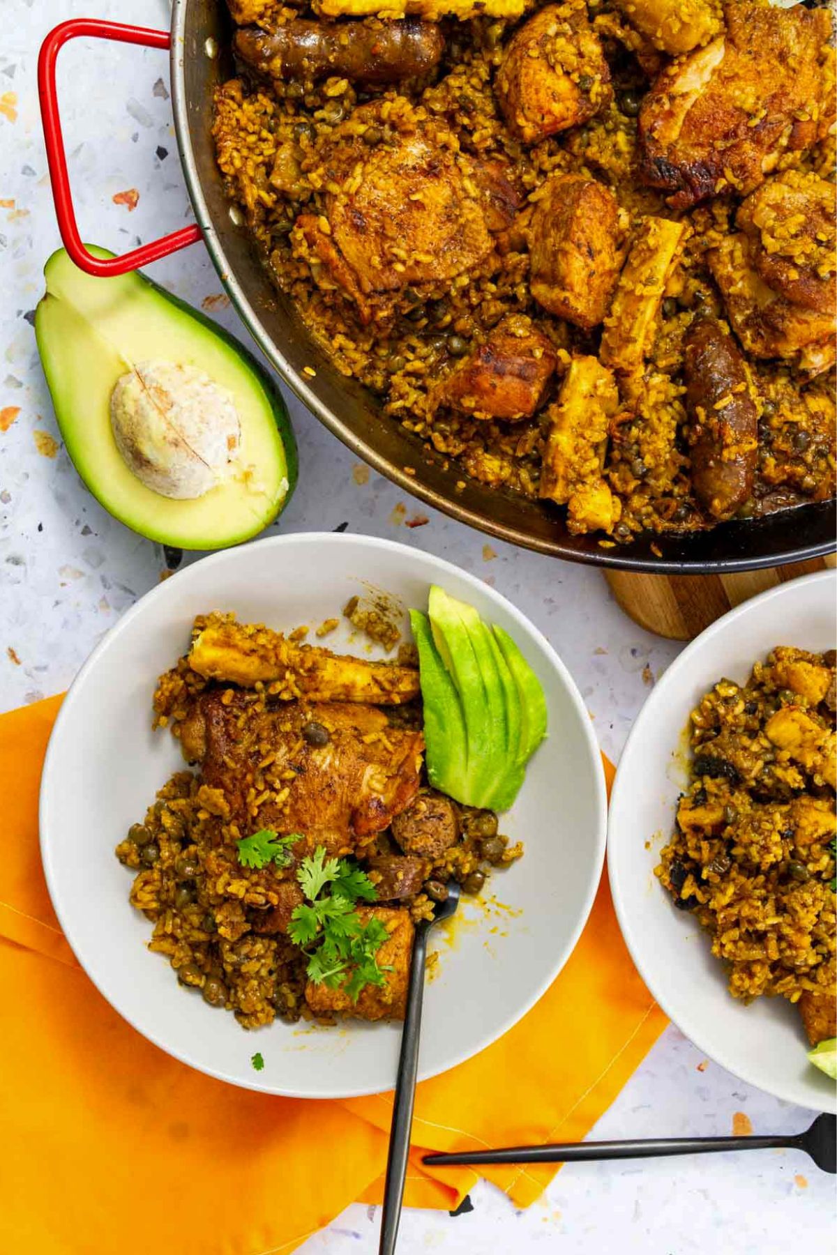 Plated Puerto Rican chicken paella with green pigeon peas, sausage, and plantains, served with sliced avocado and garnished with fresh cilantro.