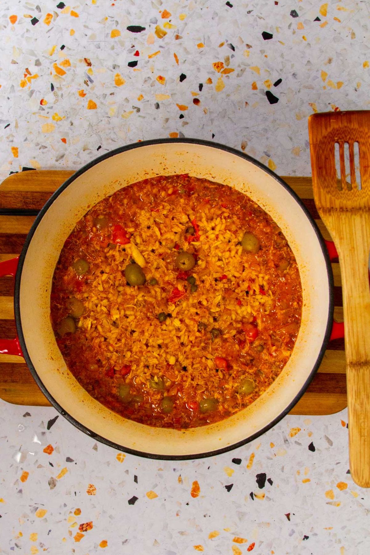 Overhead view of Puerto Rican Crab Rice (Arroz con Jueyes) in a red Dutch oven, showing rice simmered with crab meat, olives, red peppers, and sofrito. A wooden slotted spatula rests beside the pot on a terrazzo countertop.