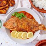 Whole Puerto Rican fried red snapper on a plate with tomato sauce, lemon slices, and plantains.
