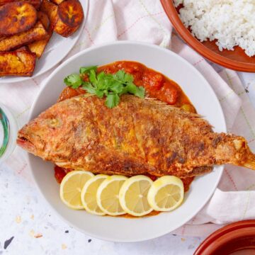 Whole Puerto Rican fried red snapper on a plate with tomato sauce, lemon slices, and plantains.
