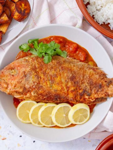 Whole Puerto Rican fried red snapper on a plate with tomato sauce, lemon slices, and plantains.