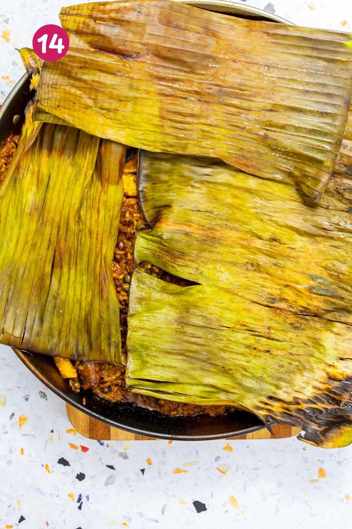 Steamed Puerto Rican paella is being covered with wilted banana leaves, peaking through bits of chicken, sausage, and rice.