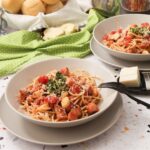 Two plates of Seafood Pomodoro pasta served with gremolata and cheese, set on a terrazzo counter with a green napkin and bread basket in the background.