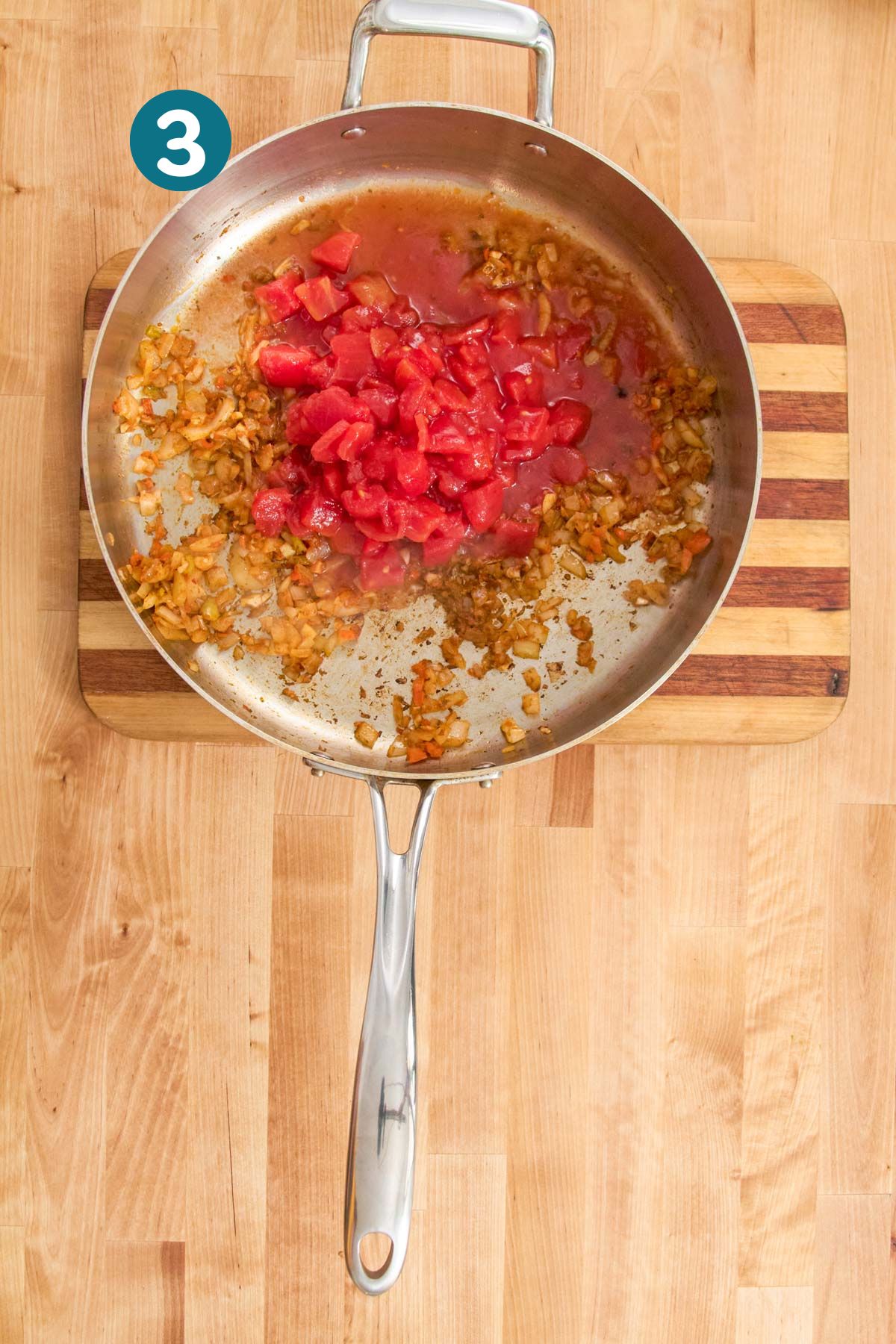 Canned diced tomatoes are added to the cooked sofrito base in a stainless steel skillet.
