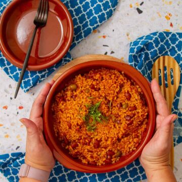 Hands holding a clay bowl of Puerto Rican arroz mamposteao, rice mixed with beans and olives, garnished with fresh herbs.