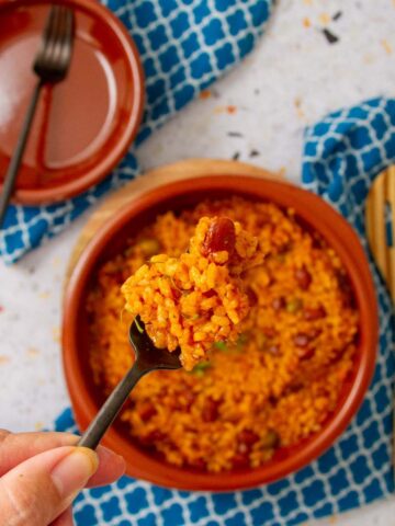 Close-up of a forkful of arroz mamposteao, Puerto Rican rice and beans, held above a bowl on a blue patterned cloth.