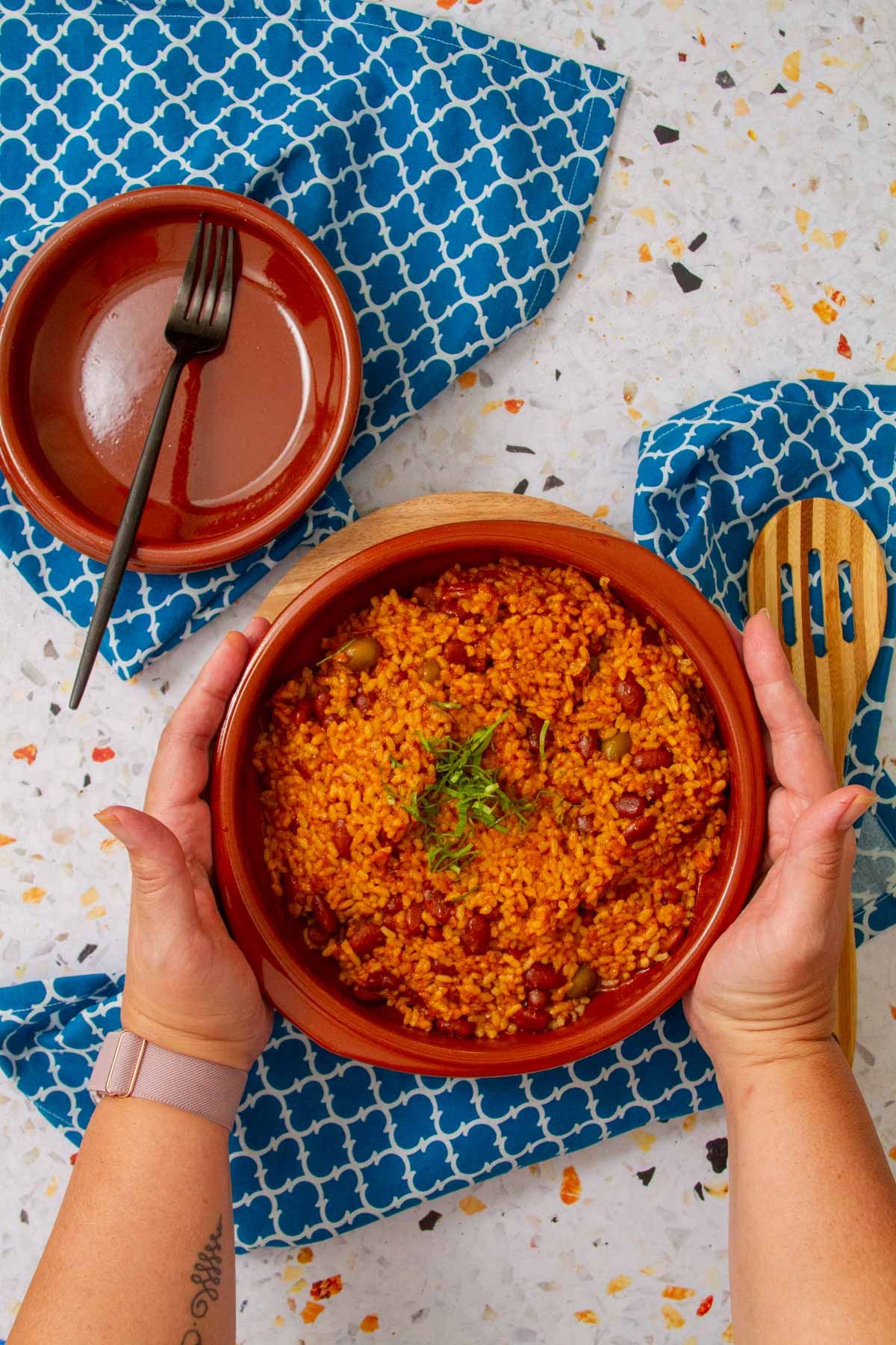 Hands holding a clay bowl filled with arroz mamposteao, garnished with fresh herbs, set on a blue patterned cloth.