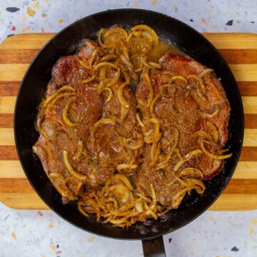 Biftec encebollado in a pan with caramelized onions and a rich pan sauce.