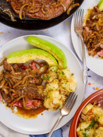 Plates of biftec encebollado with onions, avocado, and yuca served family-style.