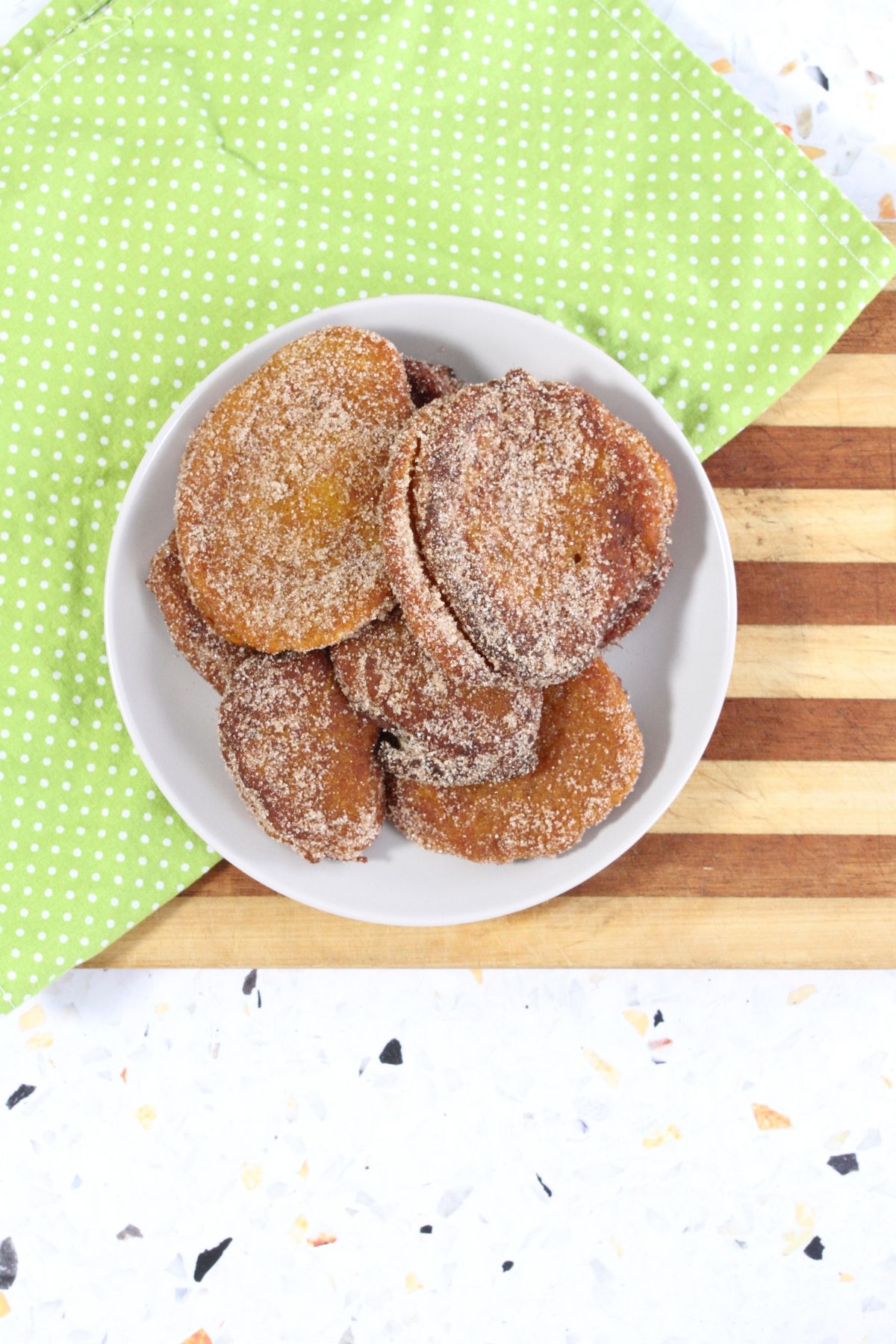 A white plate stacked with sugar-coated pumpkin fritters on a striped wooden board and a green polka-dotted napkin.