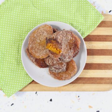 A white plate stacked with sugar-coated pumpkin fritters on a striped wooden board and a green polka-dotted napkin.